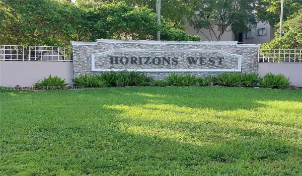 Monolithic stone-veneer community signage featuring decorative trim, bordered by a manicured lawn, established landscaping, and perimeter fencing