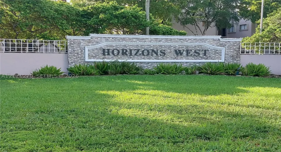 Monolithic stone-veneer community signage featuring decorative trim, bordered by a manicured lawn, established landscaping, and perimeter fencing