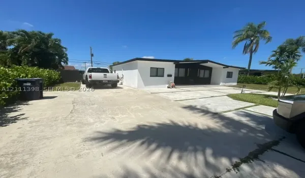Single-story exterior featuring white stucco siding, a flat roofline, an expansive concrete driveway, and custom paver sections with turf inlays