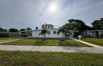 Single-story exterior featuring a painted masonry facade, flat roofline, attached covered carport, concrete walkway, and mature palm trees