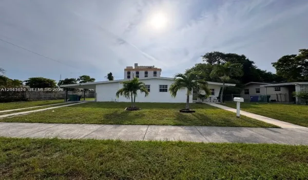 Single-story exterior featuring a painted masonry facade, flat roofline, attached covered carport, concrete walkway, and mature palm trees