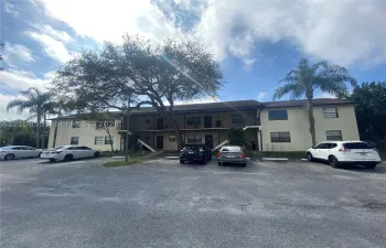 Two-story exterior featuring a neutral stucco finish, covered balcony corridors, mature shade trees, and an asphalt parking lot