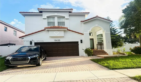 Two-story exterior featuring a light-toned stucco facade, arched entryway, clay-tile roof, and a paved driveway leading to an attached two-car garage