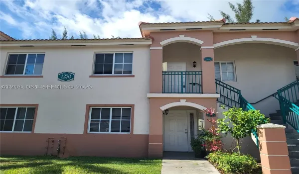 Stucco exterior featuring dual-level covered porches, terra-cotta-style roof tiles, decorative arched architectural detailing, and metal stair railing