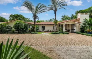 Mediterranean-style exterior featuring a clay tile roof, expansive brick-paver driveway, stucco facade, decorative wrought iron gated entry, and mature tropical landscaping