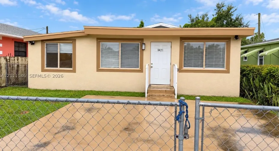 Single-story exterior featuring a light-toned stucco facade, flat roofline, concrete driveway, and white entry door with a metal handrail
