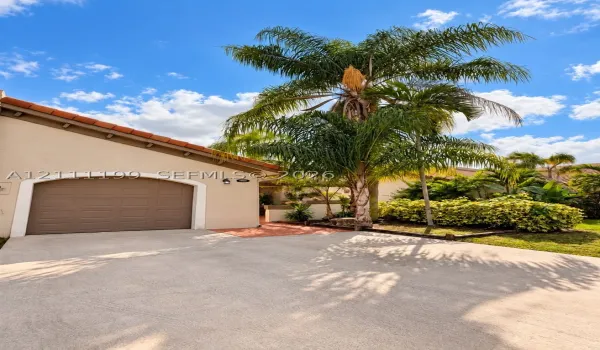 Stucco exterior featuring a concrete driveway, arched garage door, and mature palm trees with landscaped shrubbery