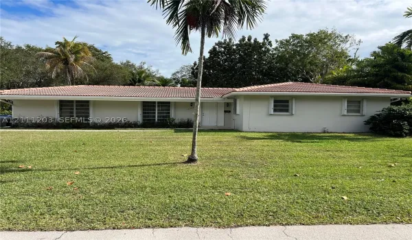 Single-story exterior featuring a light-toned stucco facade, tiled roof, horizontal jalousie windows, and a central paneled entry door, set upon a sprawling lawn with mature palm trees