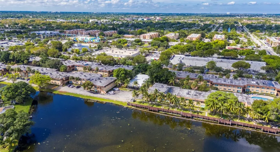 Aerial perspective featuring multi-story residential buildings, water frontage with a wooden boardwalk, mature trees, paved surface parking, and manicured landscaping