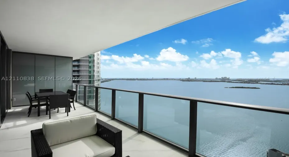 Covered balcony featuring large-format stone-finish flooring and floor-to-ceiling glass panel railings overlooking a body of water