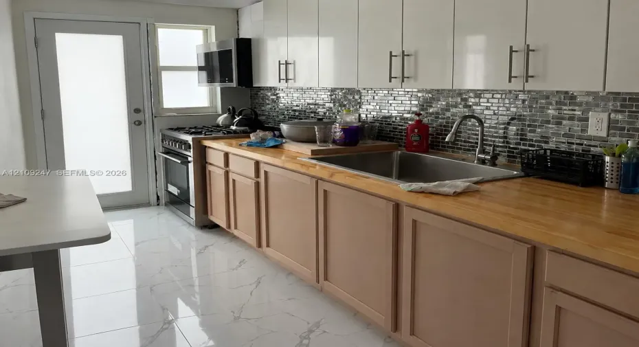 Kitchen featuring high-gloss white upper cabinetry, wood-surface countertops, mosaic tile backsplash, stainless steel appliances, and large-format porcelain tile flooring