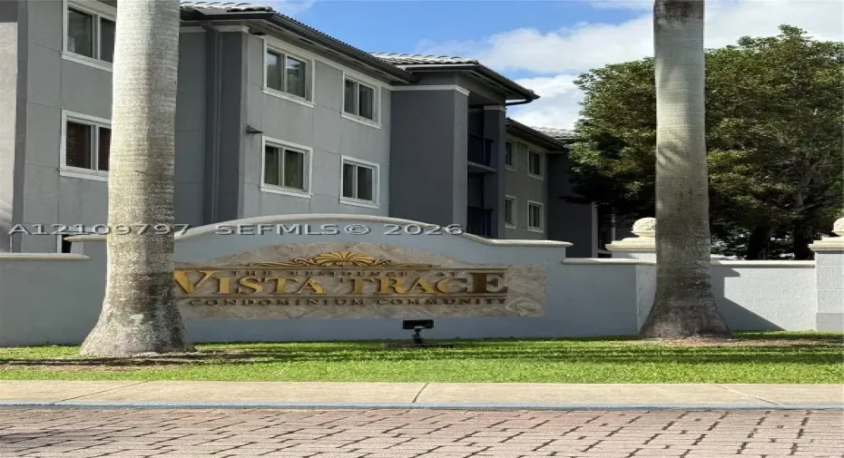 Entrance monument featuring stone veneer accenting, gold-tone lettering, and concrete pillars, set within a landscaped perimeter with mature palms and multi-story gray stucco siding