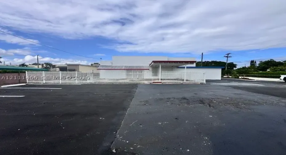 Commercial exterior featuring a paved parking lot, metal perimeter fencing, covered entryway, and white stucco facade