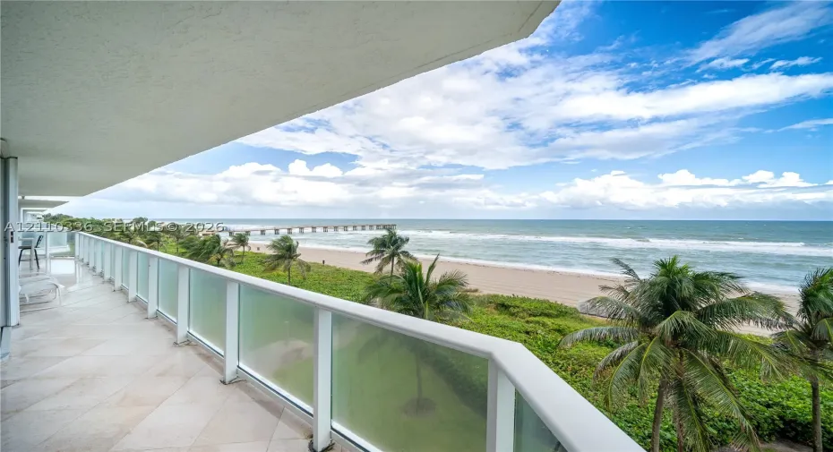 Covered balcony featuring tile flooring and a glass-paneled railing overlooking the ocean, sandy beach, and palm trees