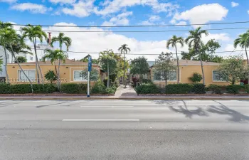 Low-rise exterior featuring a stucco facade, horizontal-sliding windows, mature palm trees, and landscaped shrubbery