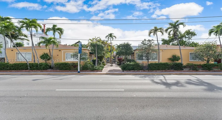 Low-rise exterior featuring a stucco facade, horizontal-sliding windows, mature palm trees, and landscaped shrubbery