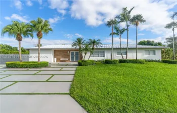 Single-story residence featuring a standing-seam metal roof, horizontal wood-panel siding, a concrete paver driveway with integrated turf, and professional landscaping with mature palm trees