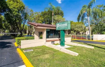 Gated community entrance featuring a guardhouse with a tiled roof, manicured landscaping, and mature palm trees