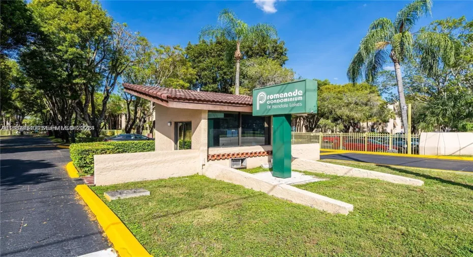 Gated community entrance featuring a guardhouse with a tiled roof, manicured landscaping, and mature palm trees