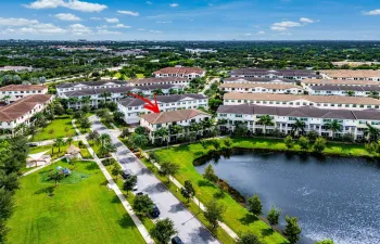 Aerial perspective featuring a multi-story residence with a clay tile roof, situated within a community offering common green spaces, palm tree landscaping, and a central water feature
