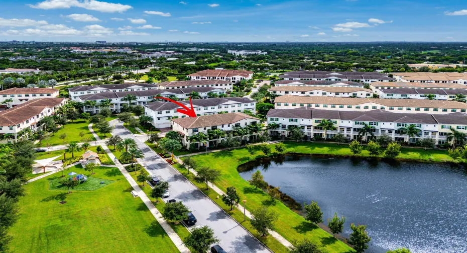 Aerial perspective featuring a multi-story residence with a clay tile roof, situated within a community offering common green spaces, palm tree landscaping, and a central water feature