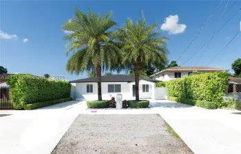 Single-story residence featuring a white exterior, hip roof, mature palm trees, and manicured privacy hedges
