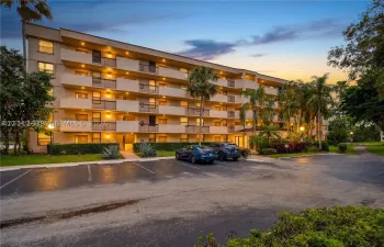 Multi-story residential building exterior featuring private balconies, asphalt parking area, and mature palm landscaping