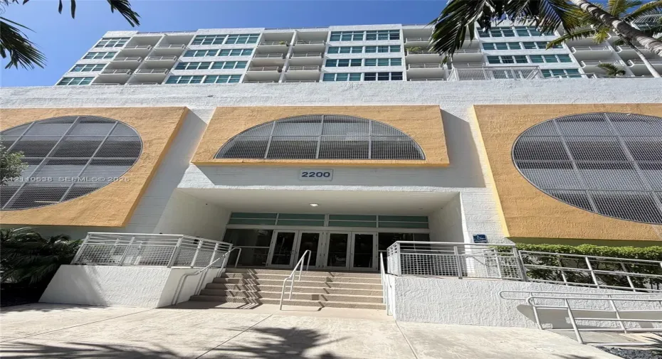 High-rise exterior featuring a white concrete facade, arched windows, tiered balconies, and a central entrance with metal railings and concrete stairs