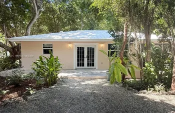 Single-story exterior featuring a metal roof, light-toned stucco facade, and double French doors, surrounded by mature trees and a gravel driveway