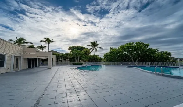 Outdoor recreation area featuring a multi-section swimming pool, expansive tiled decking, metal perimeter fencing, and mature palm trees