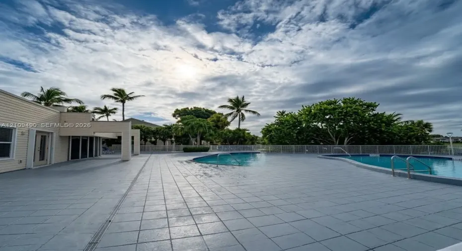 Outdoor recreation area featuring a multi-section swimming pool, expansive tiled decking, metal perimeter fencing, and mature palm trees
