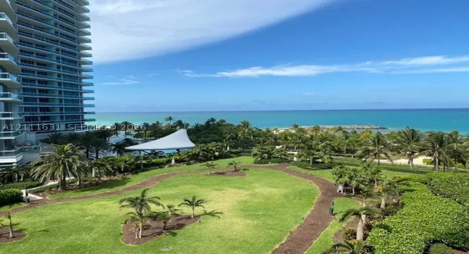Expansive grounds featuring manicured lawns, winding landscape pathways, mature palm trees, and an open-air pavilion, set against an ocean backdrop