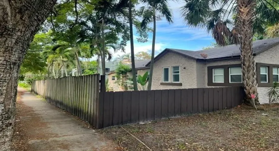 Stucco exterior featuring dark wood-tone window trim, a vertical wood privacy fence, and mature palm tree landscaping