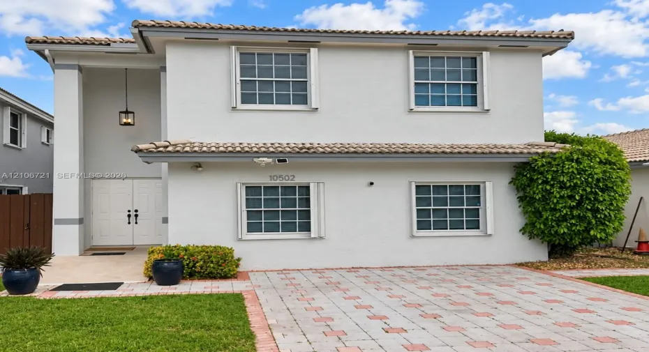 Two-story exterior featuring light-toned stucco, a concrete tile roof, double front entry doors, and a decorative paver driveway