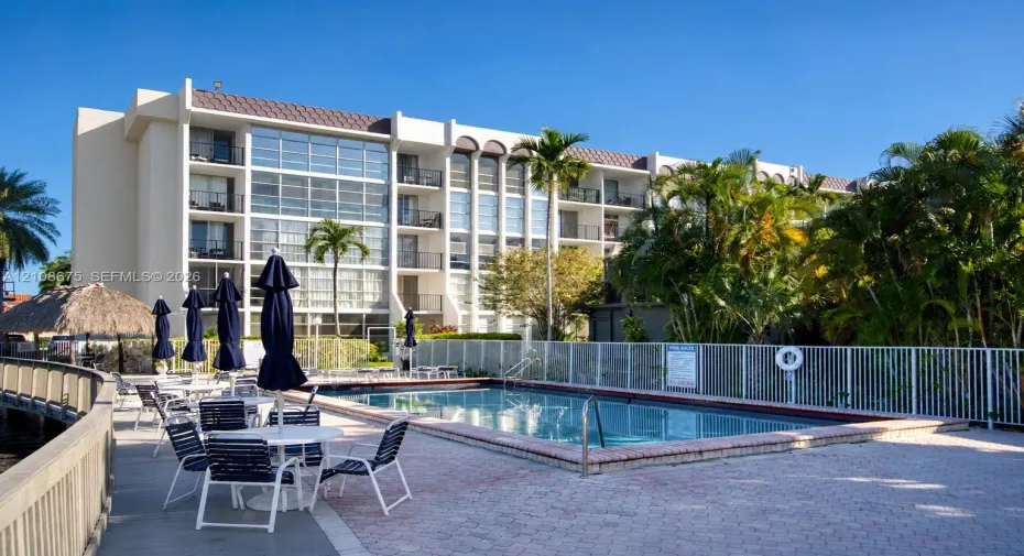 Multi-story residential building featuring a swimming pool, brick-paver pool deck, perimeter fencing, and mature palm trees