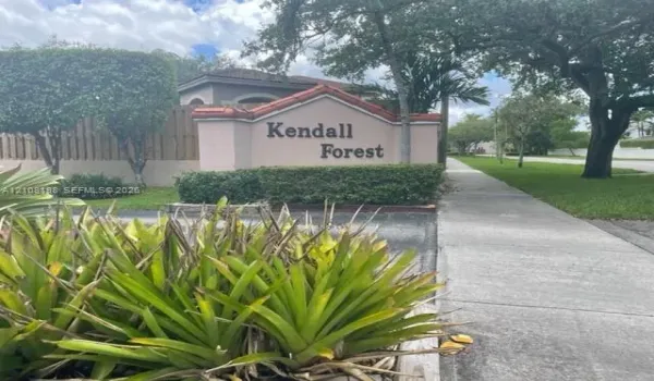 Stucco monument signage with tile detailing, manicured shrubbery, mature trees, and concrete sidewalk