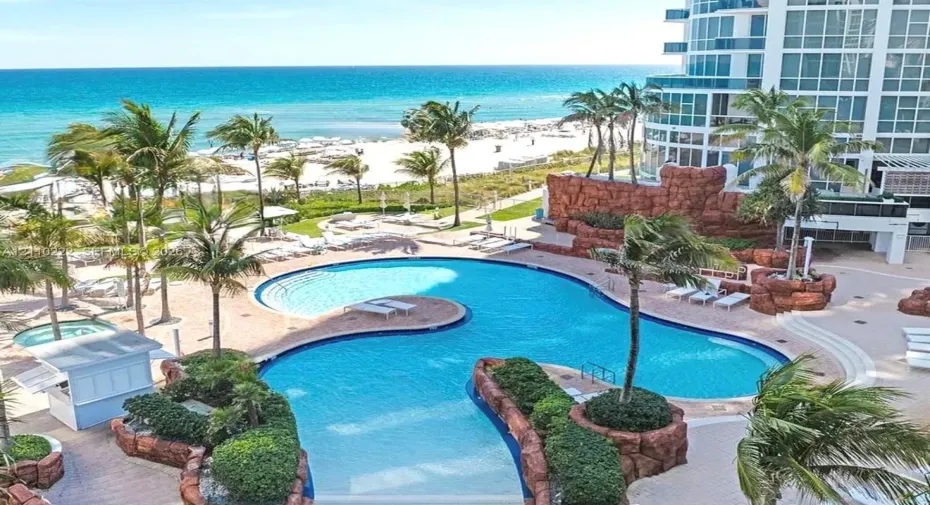 Resort-style pool area featuring tiered rock formations, expansive paved decking, lush landscaping, and integrated water features