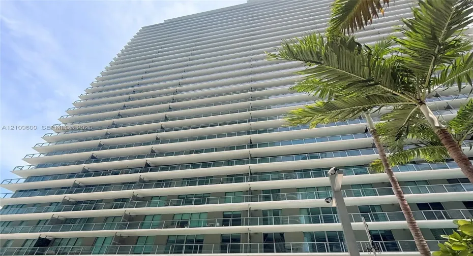 High-rise building exterior featuring floor-to-ceiling glass windows, wraparound balconies with glass railings, and structural concrete overhangs