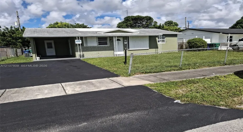 Single-story residence featuring a brick facade, covered carport, chain-link perimeter fencing, and an asphalt driveway