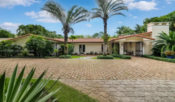 Mediterranean-style exterior featuring a clay tile roof, expansive brick-paver driveway, stucco facade, decorative wrought iron gated entry, and mature tropical landscaping