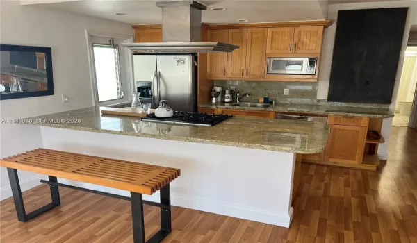 Kitchen featuring a breakfast bar with stone-surfaced countertops, wood cabinetry, stainless steel gas cooktop, stainless steel chimney-style range hood, and wood-finish flooring