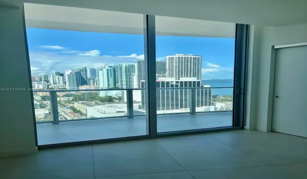 Floor-to-ceiling glass sliding doors, covered balcony with glass railing, large-format tile flooring, and urban skyline and water backdrop