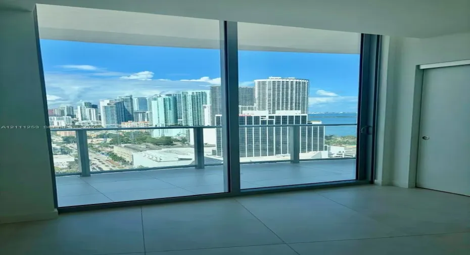 Floor-to-ceiling glass sliding doors, covered balcony with glass railing, large-format tile flooring, and urban skyline and water backdrop