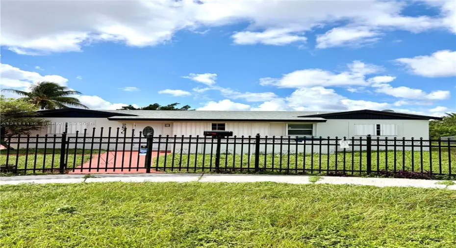 Single-story residence featuring a light-toned stucco exterior, a dark-toned gable roof, decorative window shutters, and a black wrought-iron perimeter fence