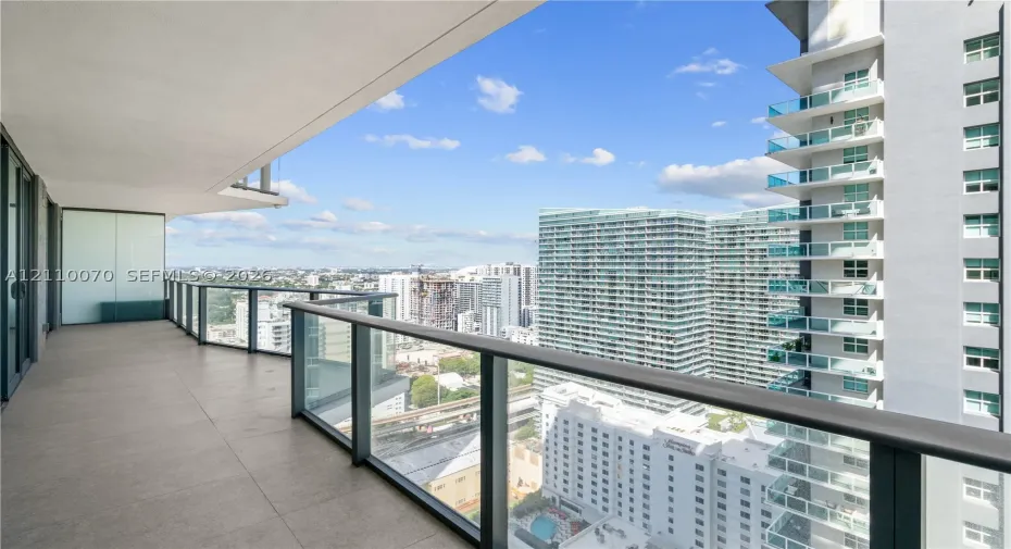 Covered balcony featuring oversized tile flooring, a glass panel railing with a dark metal frame, and privacy glass partition