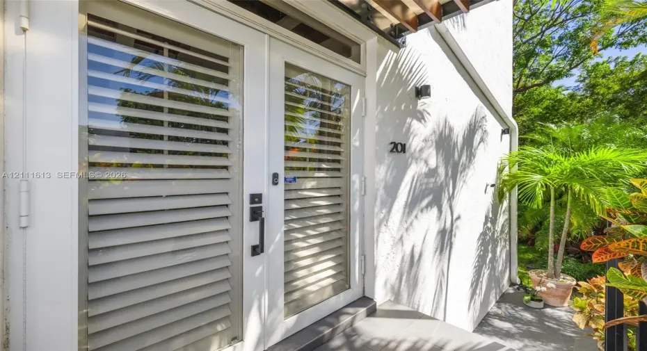 Exterior entrance featuring double glass-paneled doors with integrated horizontal louvers, a transom window, matte black hardware, and an overhead wooden pergola element