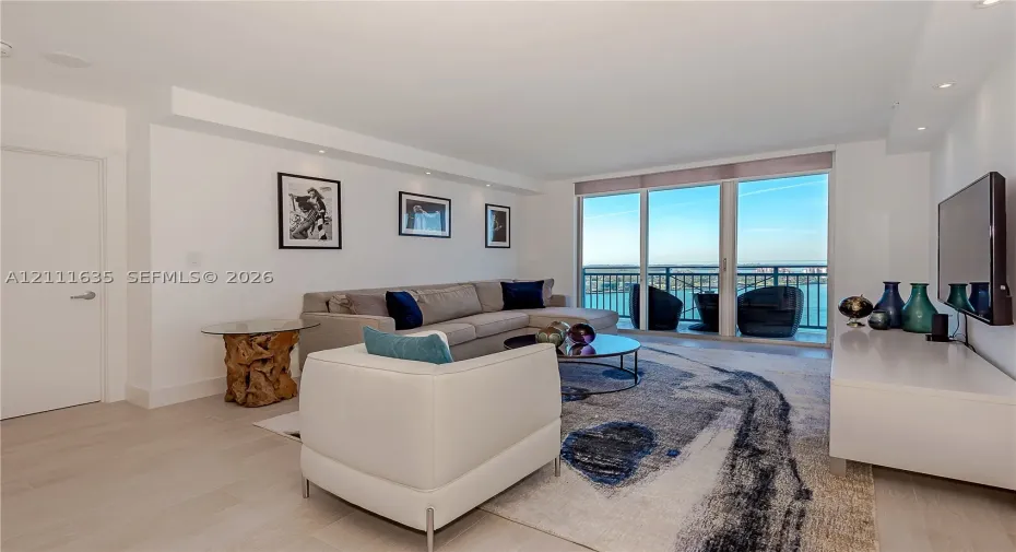 Living area featuring light-toned flooring, recessed ceiling lighting, and floor-to-ceiling glass panel doors opening to a balcony with metal railings