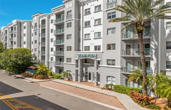 Multi-story building exterior featuring a gray and white facade, metal balconies, integrated signage, and lush palm-lined landscaping