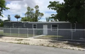 Single-story exterior featuring a gray stucco facade, flat roofline, metal window security bars, and a full-perimeter white metal picket fence