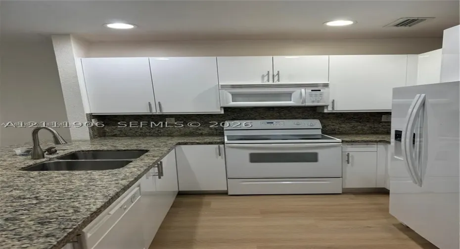 Kitchen featuring white cabinetry, stone-surface countertops, dual-basin stainless steel sink with high-arc faucet, integrated appliances, and wood-finish flooring